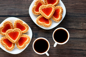 Heart shaped red jam cookies and two cups of coffee on a brown wooden background.