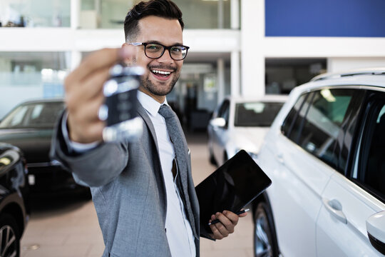 Good looking, cheerful and friendly salesman poses in a car salon or showroom. He is holding car keys while looking at camera