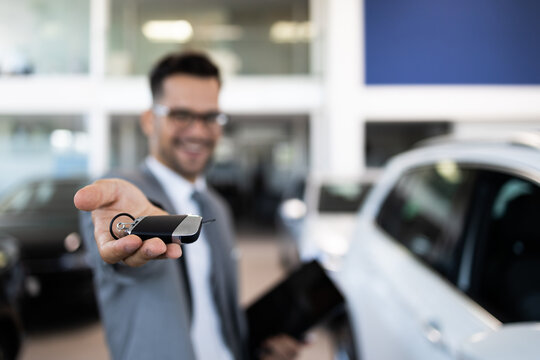 Good looking, cheerful and friendly salesman poses in a car salon or showroom. He is holding car keys while looking at camera. Selective focus on hand.