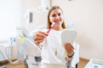 A beautiful woman in a white T-shirt sits in a dental office, looks in the mirror, smiles and holds a toothbrush in her hands. Girl with braces shows oral care. Dentistry, dental treatment