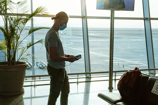 Man With Power Bank At The Airport, Charging Phone To Stay Connected During Travel, Before Flight At Departure Area. Passenger With Face Mask Traveling During Covid. Communication At Distance