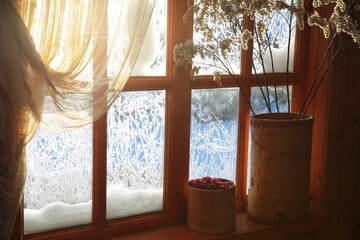 The interior of the home with red berries at the winter window with snow-covered frozen glass with sunlight on the curtain