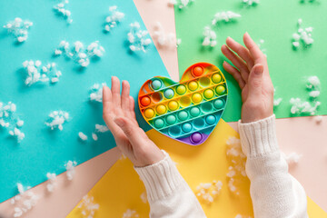 Female hands surround a silicone rainbow antistress toy in the shape of a heart on a cardboard of different colors and fluff. Trendy relaxation tool