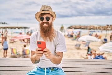 Young redhead tourist man using smartphone sitting on the bench at the beach.