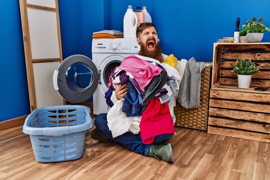 Redhead man with long beard putting dirty laundry into washing machine angry and mad screaming frustrated and furious, shouting with anger. rage and aggressive concept.