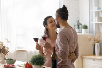 Joyful sincere loving young family couple cuddling, clinking glasses of red wine, spending romantic home date in modern kitchen, enjoying trusted candid conversation celebrating wedding anniversary.