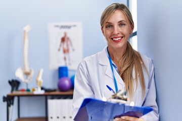 Young blonde woman wearing physiotherpist uniform writing on checklist at physiotherapy clinic