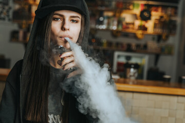 Young pretty woman in cap smoke an electronic cigarette at the vape shop. Hip-hop style. Closeup. - 480805565