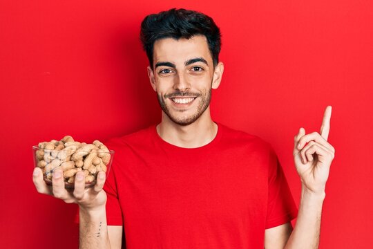 Young hispanic man holding peanuts smiling happy pointing with hand and finger to the side