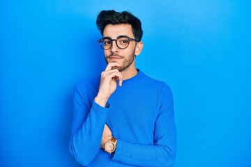 Young hispanic man wearing casual clothes and glasses looking confident at the camera with smile with crossed arms and hand raised on chin. thinking positive.