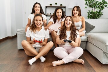 Group of young hispanic women celebrating pajamas party playing video game at home.
