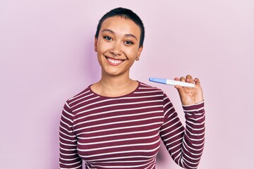 Beautiful hispanic woman with short hair holding pregnancy test result looking positive and happy standing and smiling with a confident smile showing teeth