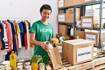 Young hispanic woman wearing volunteer uniform prepare groceries bag at charity center