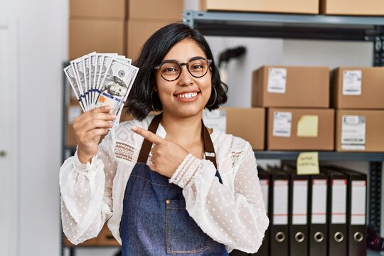 Young hispanic woman working at small business ecommerce holding dollars smiling happy pointing with hand and finger