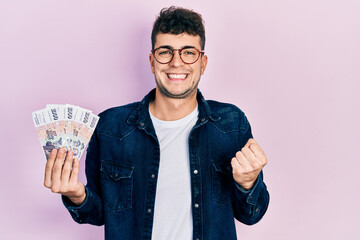 Young hispanic man holding icelandic krona banknotes screaming proud, celebrating victory and success very excited with raised arm