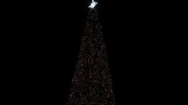 A large outdoor Christmas tree with Christmas toys and a star on top sparkles with yellow lights of garlands. New Year's paraphernalia and decorations. Moving the camera down