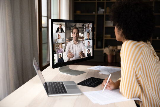 Focused young African American female employee involved in video call distant meeting with team leader and colleagues, discussing online project distantly, sitting at table in modern home office.