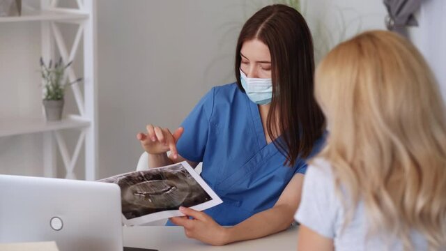 Dental problem. Doctor consultation. Medical therapy. Woman in protective mask showing teeth roentgenogram to female patient in light room interior.