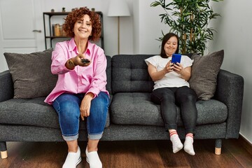 Obraz premium Mature mother and down syndrome daughter at home watching television and using smartphone