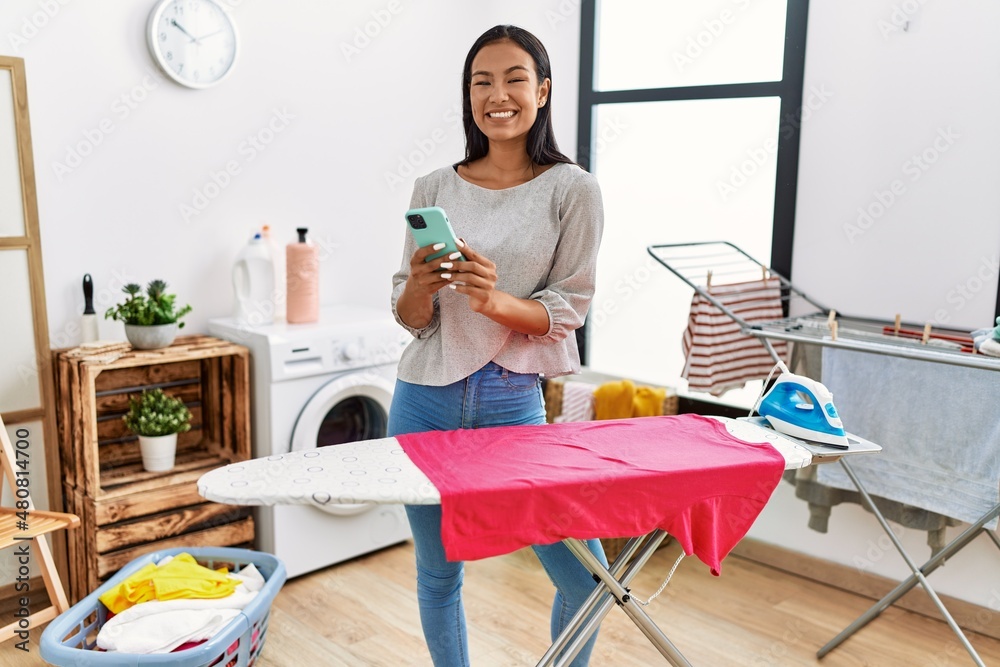 Wall mural Young latin woman using smartphone ironing clothes at laundry room