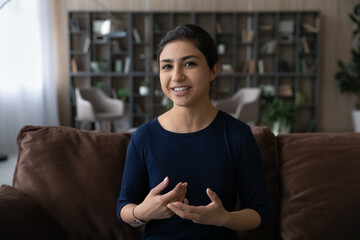 Attractive happy young Indian woman talking holding web camera call, sitting on comfortable sofa at...