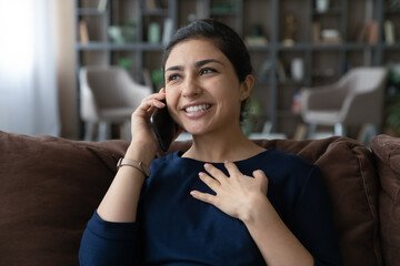 Fototapeta premium Smiling beautiful young Indian ethnic woman talking enjoying pleasant cellphone conversation, calling friends or sharing life news with family resting on cozy sofa, distant communication concept.