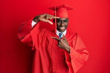 Young african american man wearing graduation cap and ceremony robe smiling making frame with hands...