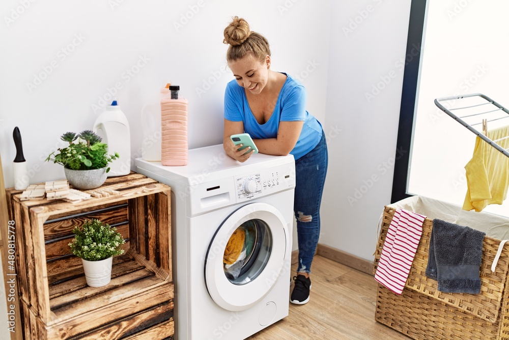 Poster young blonde girl using smartphone waiting for washing machine at laundry room