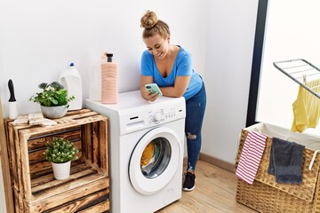 Young blonde girl using smartphone waiting for washing machine at laundry room
