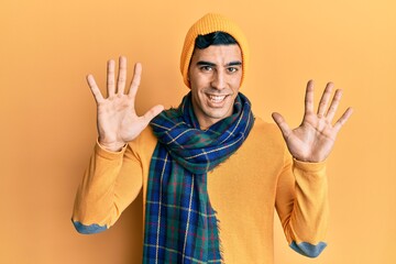 Naklejka premium Handsome hispanic man wearing wool hat and winter scarf showing and pointing up with fingers number ten while smiling confident and happy.