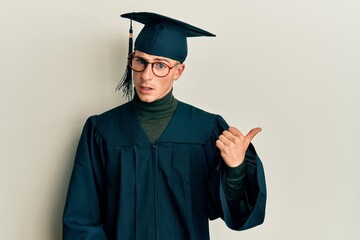 Young caucasian man wearing graduation cap and ceremony robe smiling with happy face looking and pointing to the side with thumb up.