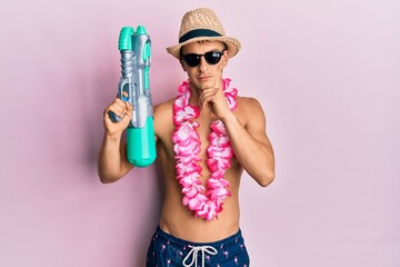 Young caucasian man wearing swimsuit and hawaiian lei holding watergun serious face thinking about...