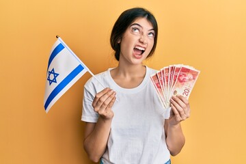 Young caucasian woman holding israel flag and shekels banknotes angry and mad screaming frustrated...