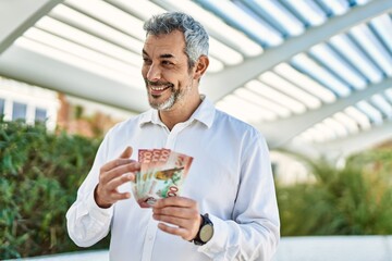 Middle age grey-haired man smiling happy counting new zealand dollars at the city.
