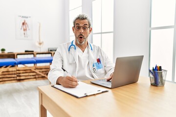 Middle age hispanic therapist man working at pain recovery clinic with laptop scared and amazed with open mouth for surprise, disbelief face