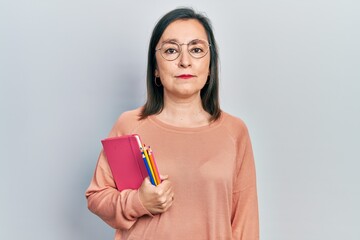 Middle age hispanic woman holding book and color pencils thinking attitude and sober expression looking self confident