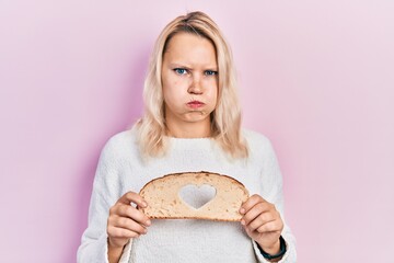Beautiful caucasian blonde woman holding bread loaf with heart shape puffing cheeks with funny...