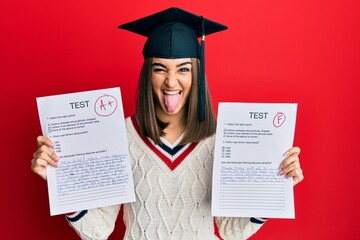 Young brunette girl wearing graduation cap showing exams sticking tongue out happy with funny...