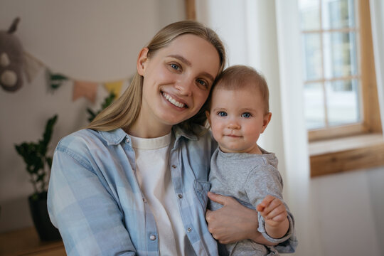 Young smiling mother holding little baby looking at camera. Beautiful family communication online, having video call. Portrait of happy influencer with cute child recording video 