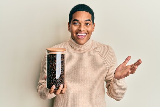 Young handsome hispanic man holding jar with coffee beans celebrating achievement with happy smile and winner expression with raised hand