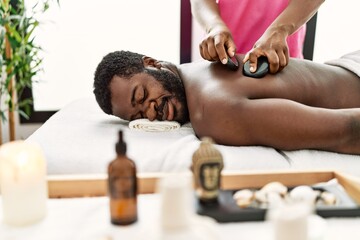 Obraz na płótnie Canvas African american man reciving back massage with black stones at beauty center.