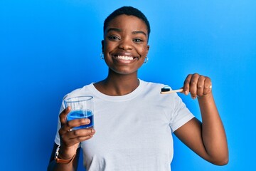 Young african american woman holding glass of mouthwash and toothbrush for fresh breath smiling...