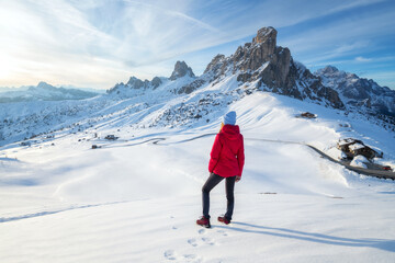 Fototapeta na wymiar Young woman in red jacket in snowy mountains at sunset in winter. Landscape with beautiful girl on the hill in snow, rocks, blue sky at sunny day. Mountain pass in Dolomites, Italy. Tourism. Travel