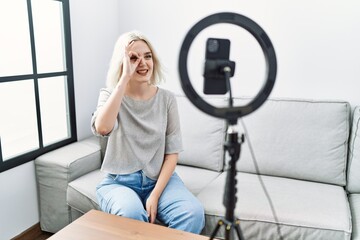 Fototapeta na wymiar Young caucasian woman recording vlog tutorial with smartphone at home doing ok gesture with hand smiling, eye looking through fingers with happy face.