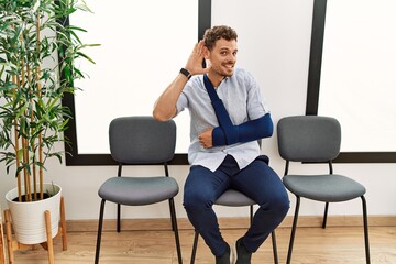 Handsome young man sitting at doctor waiting room with arm injury smiling with hand over ear listening an hearing to rumor or gossip. deafness concept.