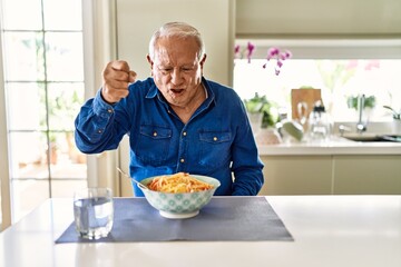 Senior man with grey hair eating pasta spaghetti at home angry and mad raising fist frustrated and...