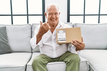 Senior man holding delivery package at home pointing thumb up to the side smiling happy with open...