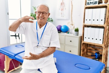 Obraz premium Senior physiotherapy man working at pain recovery clinic gesturing with hands showing big and large size sign, measure symbol. smiling looking at the camera. measuring concept.