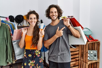 Young hispanic couple holding shopping bags at retail shop cheerful with a smile on face pointing with hand and finger up to the side with happy and natural expression