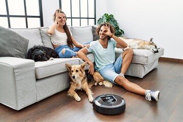 Young hispanic couple relaxing at home by vacuum robot smiling happy doing ok sign with hand on eye looking through fingers